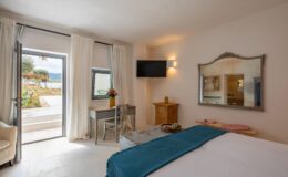Cozy bedroom interior featuring a comfortable bed with a turquoise throw, a small wooden desk with a chair, and a vase of flowers. Natural light streams in through large windows draped with white curtains, offering a view of an outdoor garden area and palm trees. A TV mounted on the wall and a vintage mirror complete the decor, enhancing the serene atmosphere.