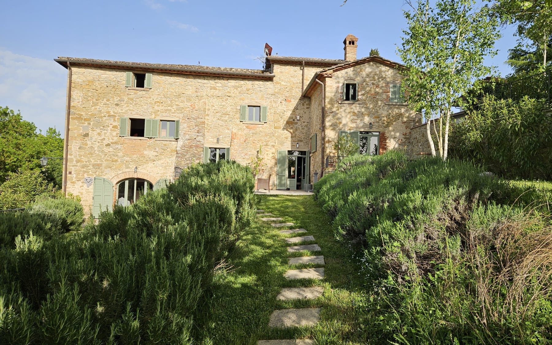 Charming rustic stone house surrounded by lush greenery and lavender, featuring a pathway leading to the entrance and a clear blue sky overhead. Ideal for a tranquil getaway in the countryside. | Only on SopranoVillas