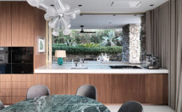 Modern kitchen interior featuring a marble dining table, sleek wooden cabinetry, and a stylish chandelier. Large windows open to a landscaped outdoor area with plants, creating a seamless indoor-outdoor living space. The kitchen includes high-end appliances and a contemporary design.