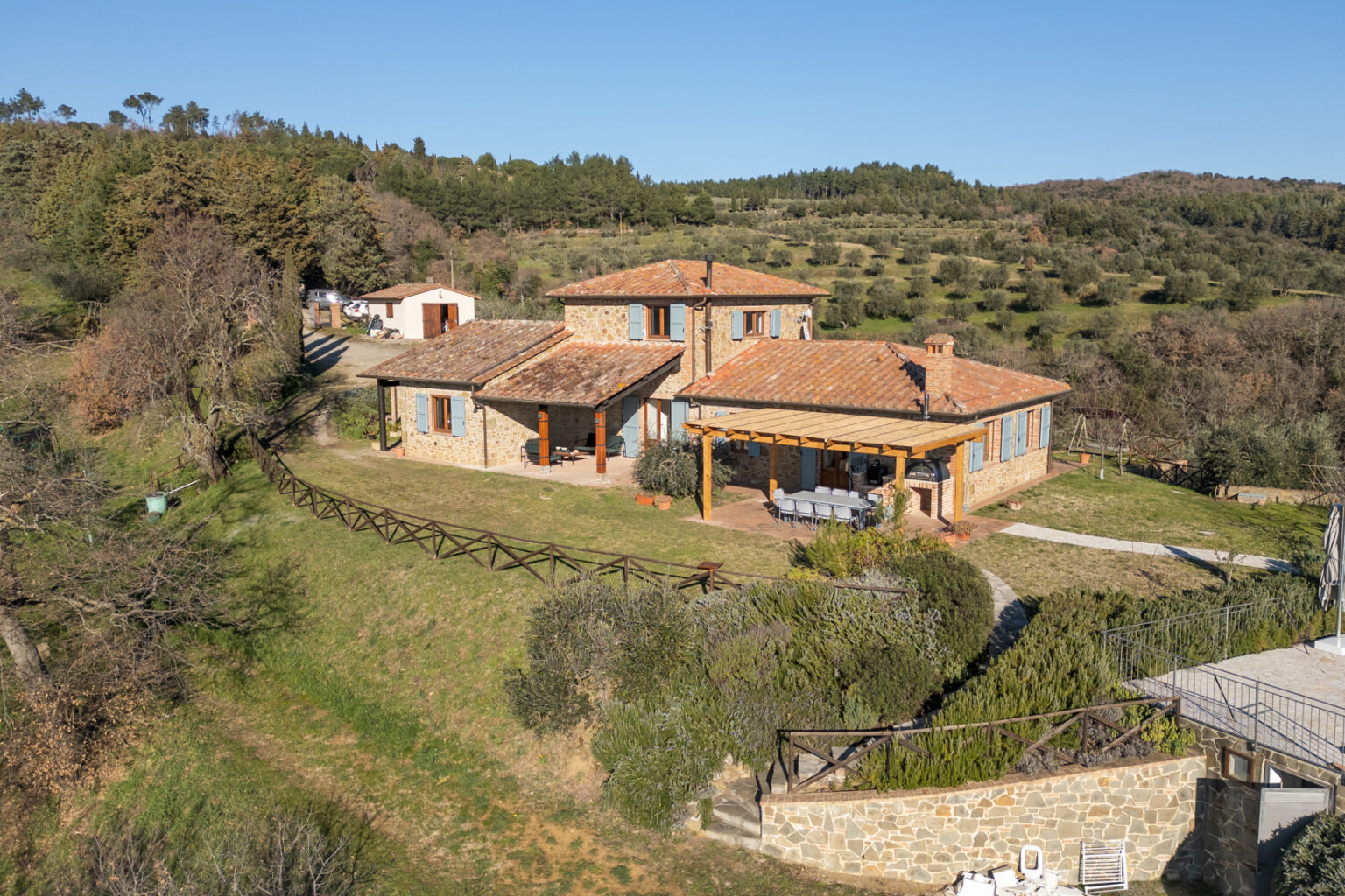 Aerial view of a charming stone villa with a terracotta roof, surrounded by lush greenery and rolling hills. The property features a spacious outdoor dining area and a well-maintained garden, showcasing the tranquility of rural living. | Only on SopranoVillas