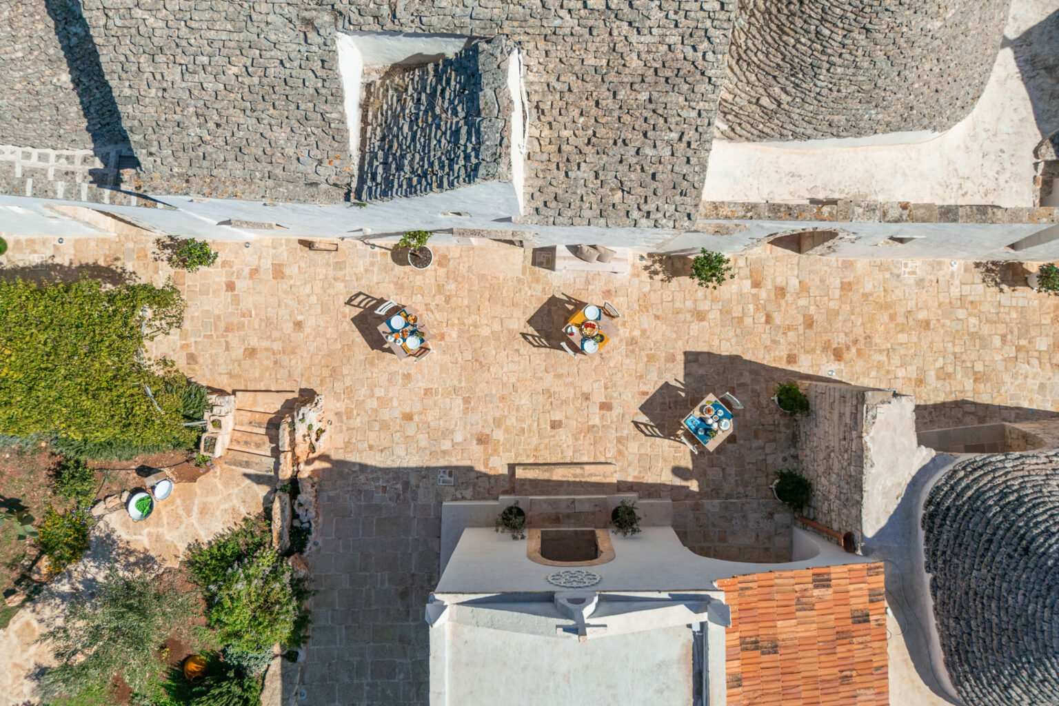 Aerial view of a charming stone courtyard surrounded by traditional trulli buildings, featuring several small tables set with colorful dishes and surrounded by lush greenery. The warm stone tiles and natural light create an inviting atmosphere for outdoor dining. | Only on SopranoVillas