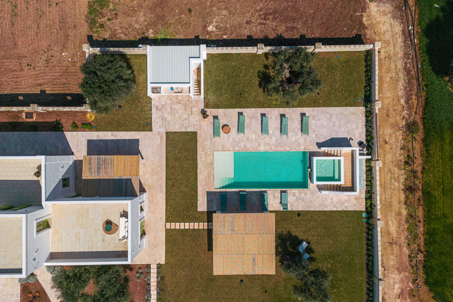 Aerial view of a luxurious modern villa featuring a swimming pool, spa area, and landscaped outdoor spaces with lounge chairs and a wooden deck, surrounded by greenery and farmland. | Only on SopranoVillas