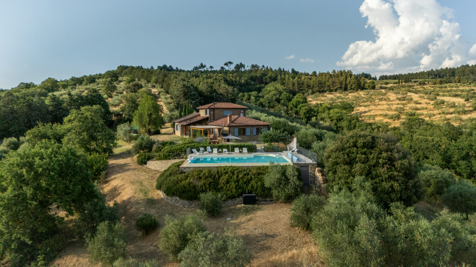 Aerial view of a rustic villa surrounded by lush greenery, featuring a modern swimming pool and landscaped gardens, located in a serene countryside setting with rolling hills and trees. Perfect for vacation rentals and nature retreats. | Only on SopranoVillas
