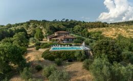 Aerial view of a rustic villa surrounded by lush greenery, featuring a modern swimming pool and landscaped gardens, located in a serene countryside setting with rolling hills and trees. Perfect for vacation rentals and nature retreats.