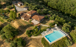 Aerial view of a rustic villa with terracotta roofs, nestled in a lush, green landscape featuring a swimming pool and outdoor seating area, surrounded by olive trees and gravel pathways.