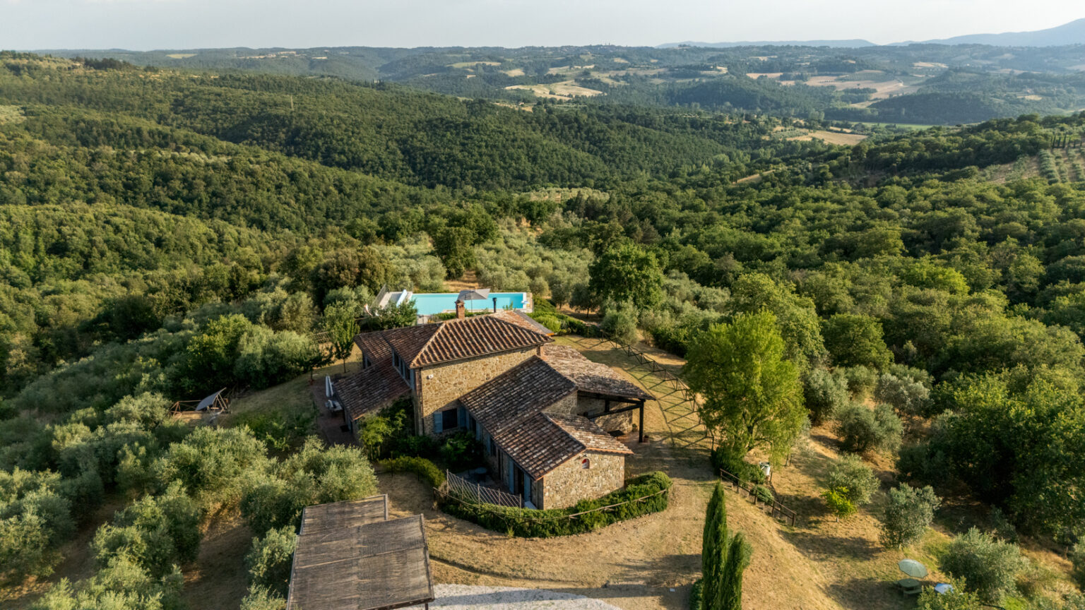 Aerial view of a charming rustic villa surrounded by lush green hills and olive groves, featuring a swimming pool and spacious outdoor areas, ideal for relaxation and nature retreats. | Only on SopranoVillas