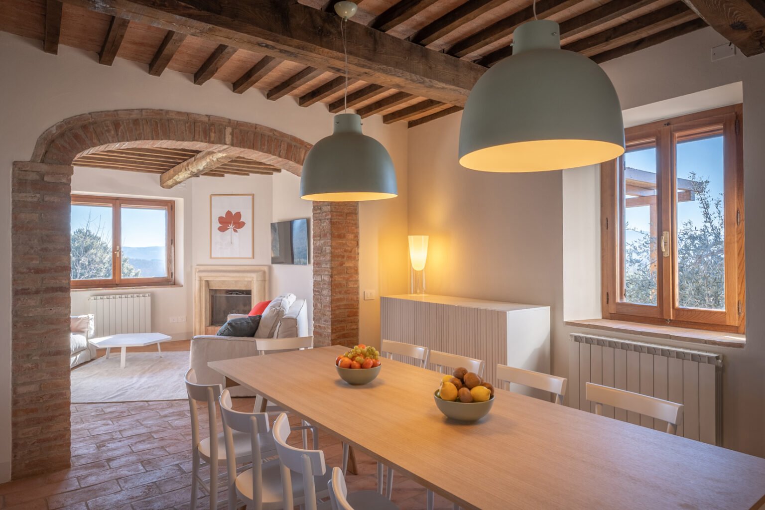 Modern dining area featuring a wooden table with bowls of fresh fruit, surrounded by white chairs. The room showcases rustic brick arches, wooden beams, and large windows that offer scenic views. Soft lighting from stylish lamps adds warmth to the inviting space. | Only on SopranoVillas