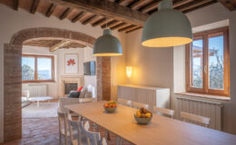 Modern dining area featuring a wooden table with bowls of fresh fruit, surrounded by white chairs. The room showcases rustic brick arches, wooden beams, and large windows that offer scenic views. Soft lighting from stylish lamps adds warmth to the inviting space.