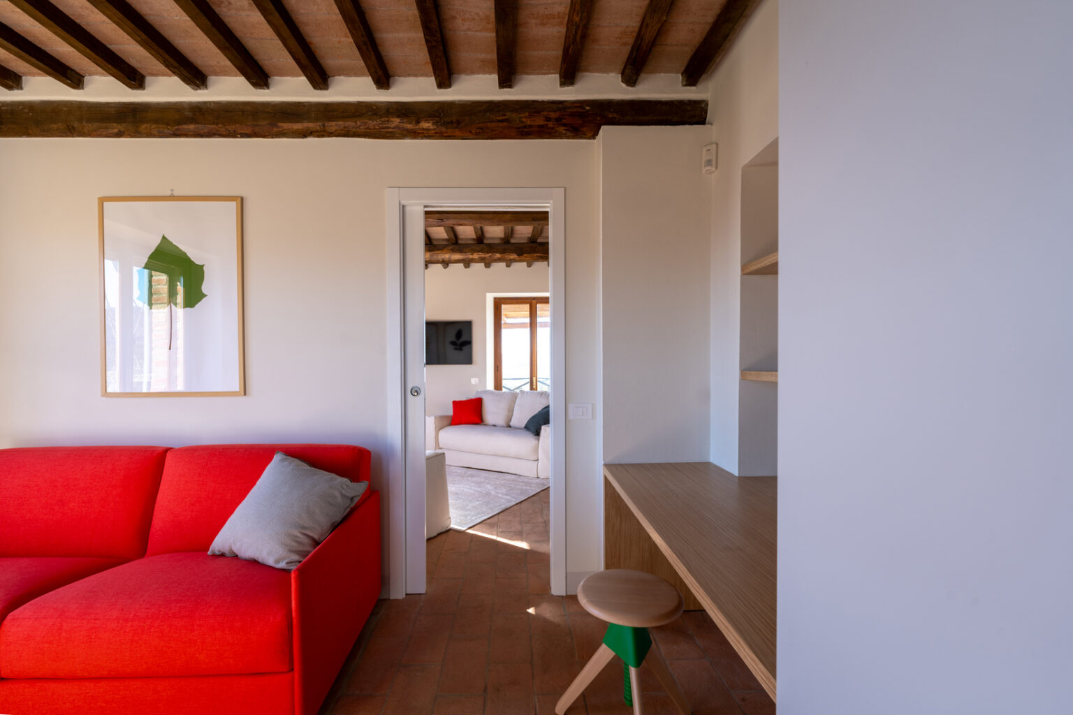 Modern living room interior featuring a vibrant red sofa with a gray pillow, wooden beams, and a minimalistic desk. A doorway leads to a cozy second seating area with natural light and simple decor, enhancing the open space feel. | Only on SopranoVillas