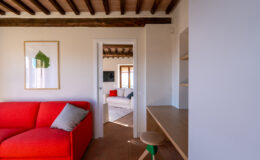 Modern living room interior featuring a vibrant red sofa with a gray pillow, wooden beams, and a minimalistic desk. A doorway leads to a cozy second seating area with natural light and simple decor, enhancing the open space feel.