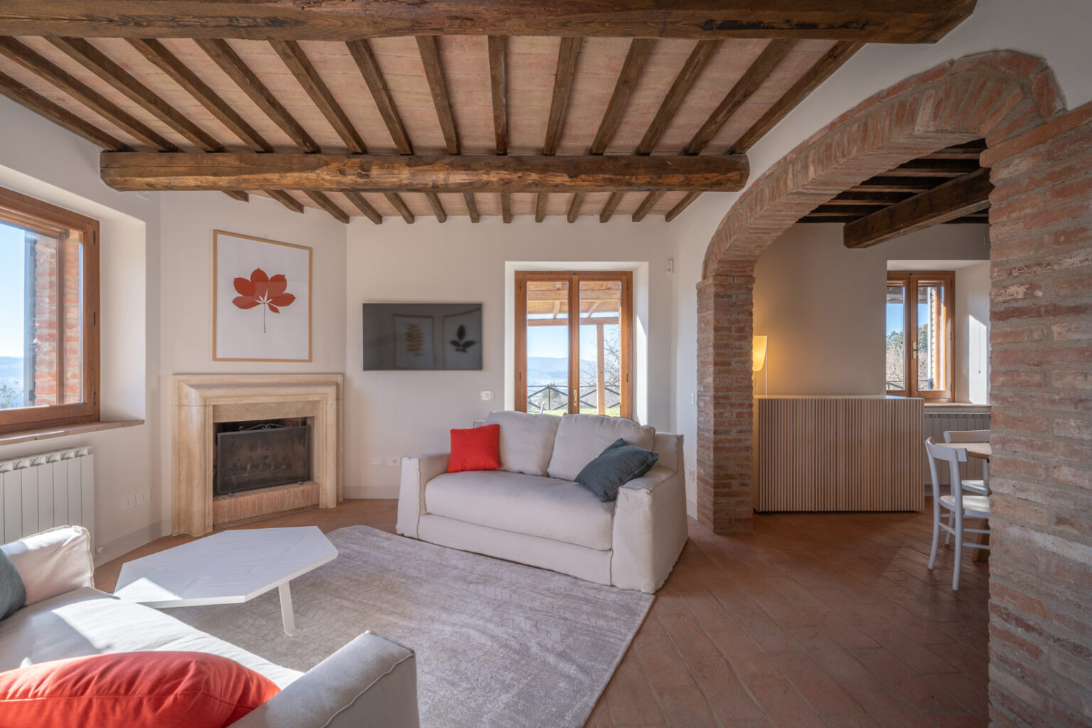 Cozy living room featuring rustic wooden beams, a stone fireplace, and large windows with scenic views. The space includes a light-colored sofa with colorful cushions, a modern coffee table, and a TV mounted on the wall, creating a warm and inviting atmosphere. | Only on SopranoVillas