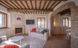 Cozy living room featuring rustic wooden beams, a stone fireplace, and large windows with scenic views. The space includes a light-colored sofa with colorful cushions, a modern coffee table, and a TV mounted on the wall, creating a warm and inviting atmosphere.