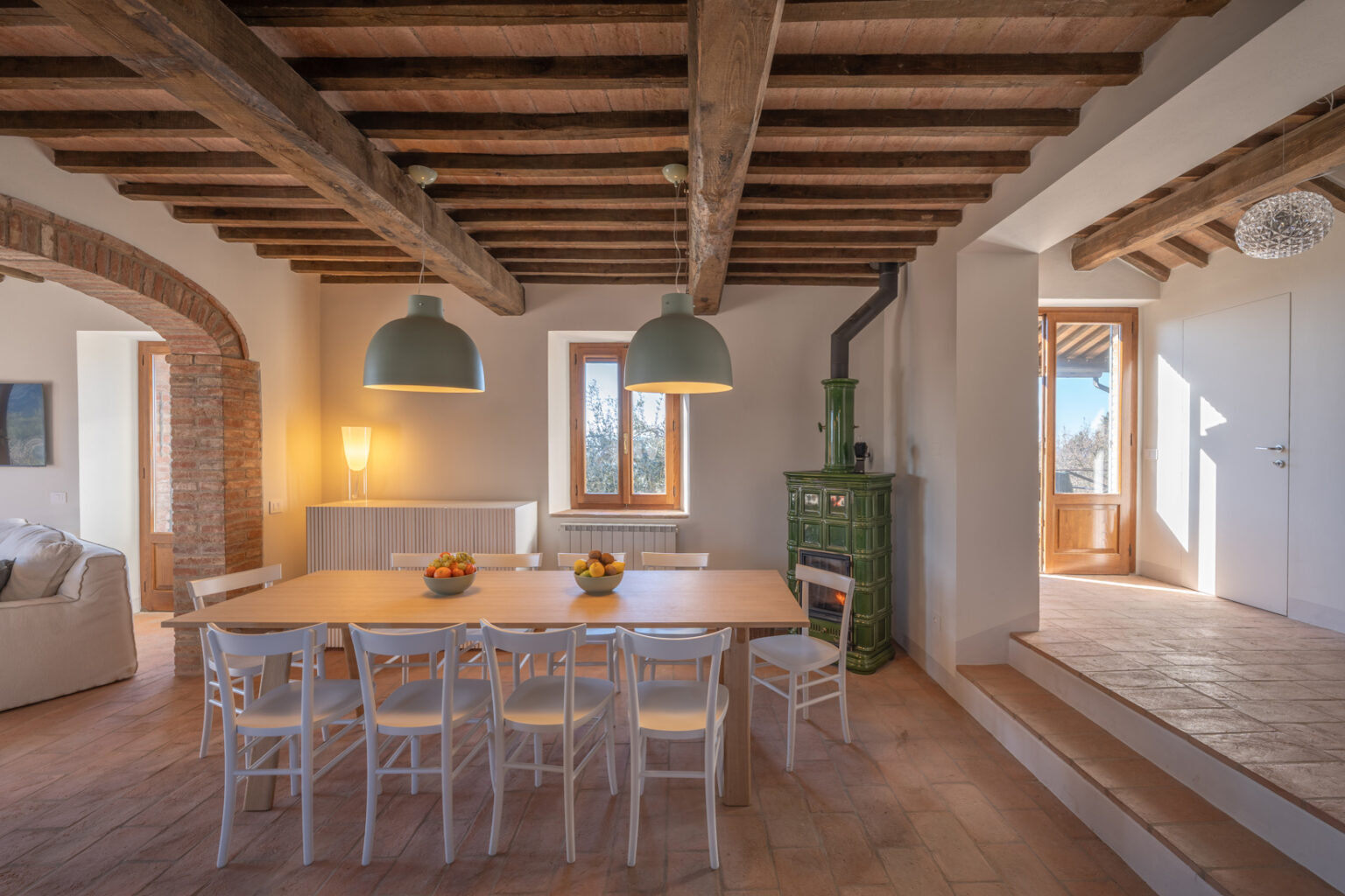 Bright and airy dining area featuring a wooden table surrounded by white chairs, complemented by green pendant lights. The space showcases rustic wooden beams and a charming green stove, with large windows allowing natural light to illuminate the room. A bowl of fresh fruits adds a touch of color to the dining table, enhancing the inviting atmosphere. | Only on SopranoVillas
