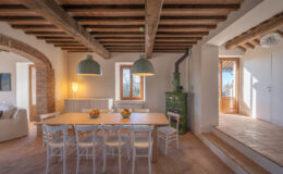 Bright and airy dining area featuring a wooden table surrounded by white chairs, complemented by green pendant lights. The space showcases rustic wooden beams and a charming green stove, with large windows allowing natural light to illuminate the room. A bowl of fresh fruits adds a touch of color to the dining table, enhancing the inviting atmosphere.
