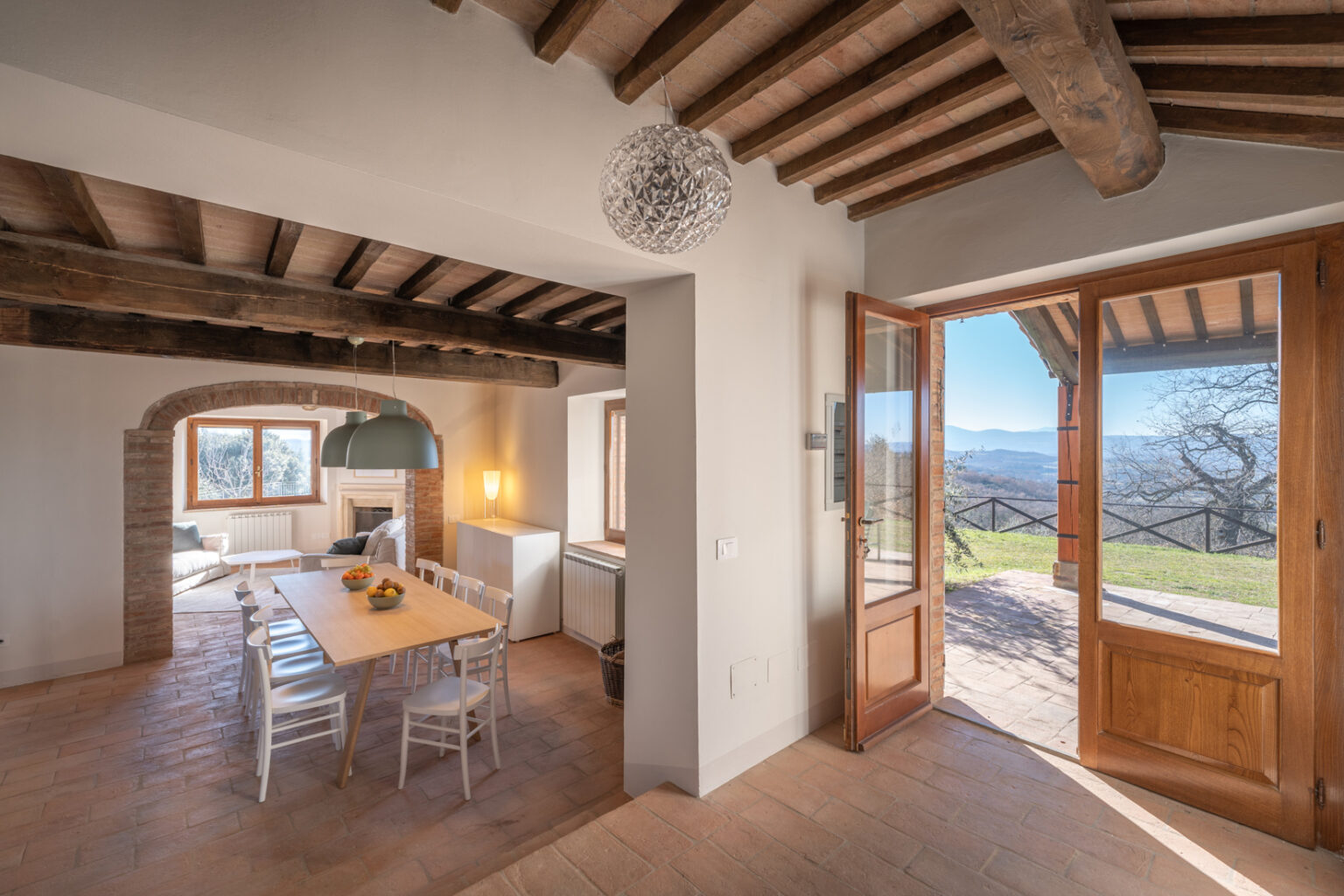 Modern rustic interior of a cozy dining area with a wooden table and white chairs, featuring exposed wooden beams and a large window. The open door leads to a scenic outdoor view, enhancing the inviting atmosphere of the space. | Only on SopranoVillas