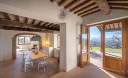 Modern rustic interior of a cozy dining area with a wooden table and white chairs, featuring exposed wooden beams and a large window. The open door leads to a scenic outdoor view, enhancing the inviting atmosphere of the space.