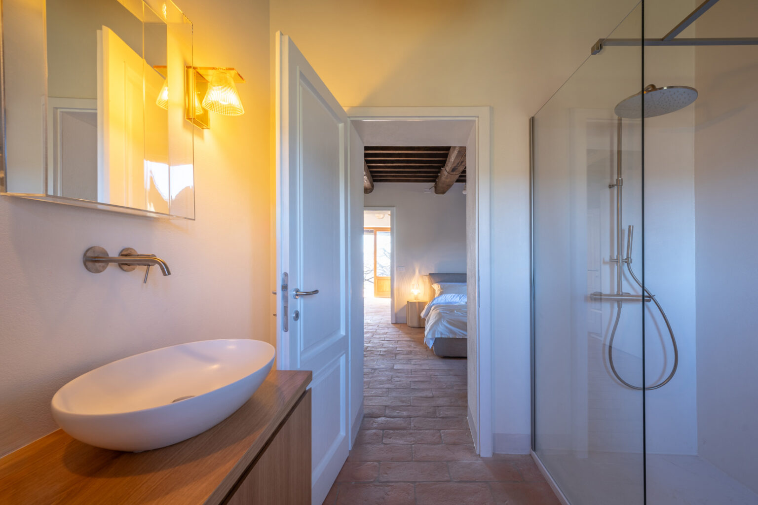Modern bathroom with a sleek vessel sink on a wooden vanity, illuminated by stylish wall sconces. The doorway leads to a serene bedroom featuring natural light and rustic wooden beams, enhancing the inviting atmosphere of the space. | Only on SopranoVillas