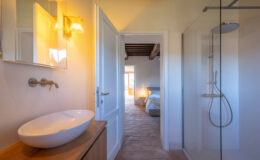 Modern bathroom with a sleek vessel sink on a wooden vanity, illuminated by stylish wall sconces. The doorway leads to a serene bedroom featuring natural light and rustic wooden beams, enhancing the inviting atmosphere of the space.