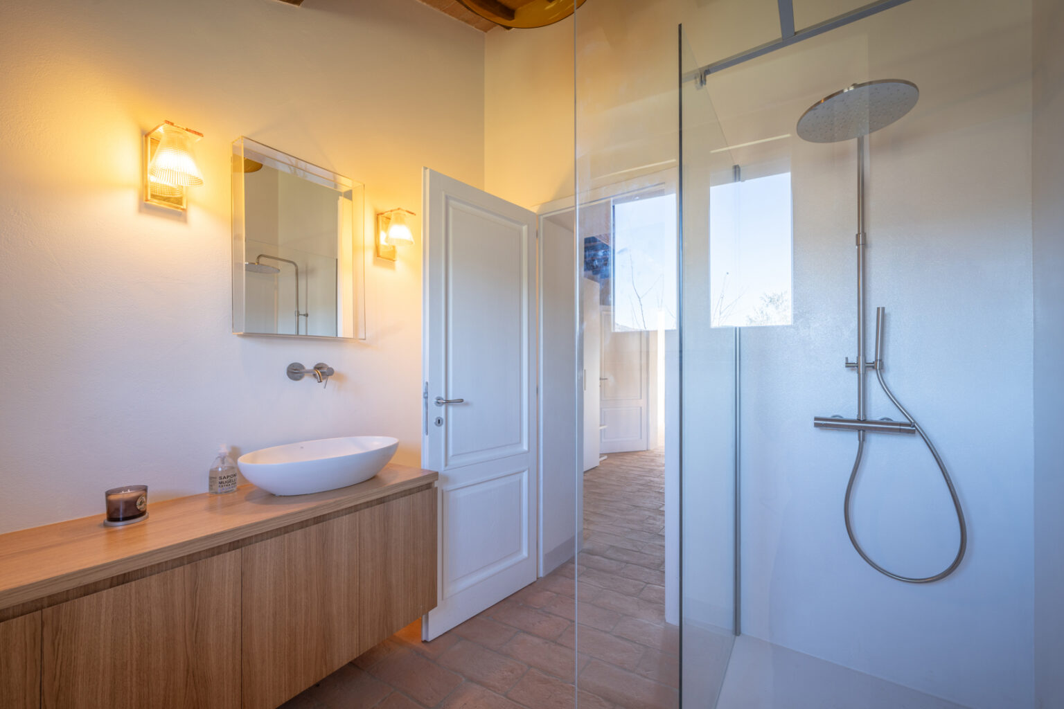 Modern bathroom interior featuring a stylish shower with glass enclosure, a sleek white vessel sink on a wooden vanity, and warm lighting fixtures. The design emphasizes minimalism and natural materials, creating an inviting and serene atmosphere. | Only on SopranoVillas