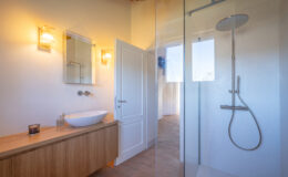 Modern bathroom interior featuring a stylish shower with glass enclosure, a sleek white vessel sink on a wooden vanity, and warm lighting fixtures. The design emphasizes minimalism and natural materials, creating an inviting and serene atmosphere.
