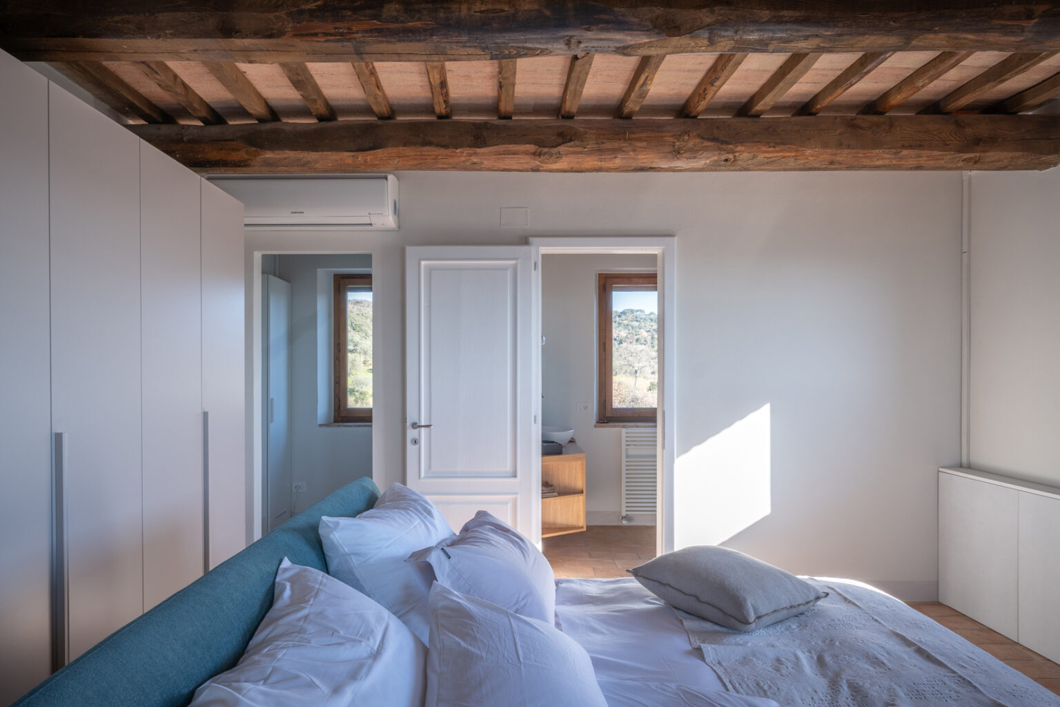 Modern bedroom interior featuring a cozy bed with white linens and a decorative pillow, wooden beams on the ceiling, and natural light streaming through large windows. The room includes a built-in wardrobe and a glimpse of an adjoining room with a wooden cabinet. Ideal for relaxation and comfort. | Only on SopranoVillas