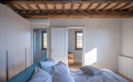 Modern bedroom interior featuring a cozy bed with white linens and a decorative pillow, wooden beams on the ceiling, and natural light streaming through large windows. The room includes a built-in wardrobe and a glimpse of an adjoining room with a wooden cabinet. Ideal for relaxation and comfort.