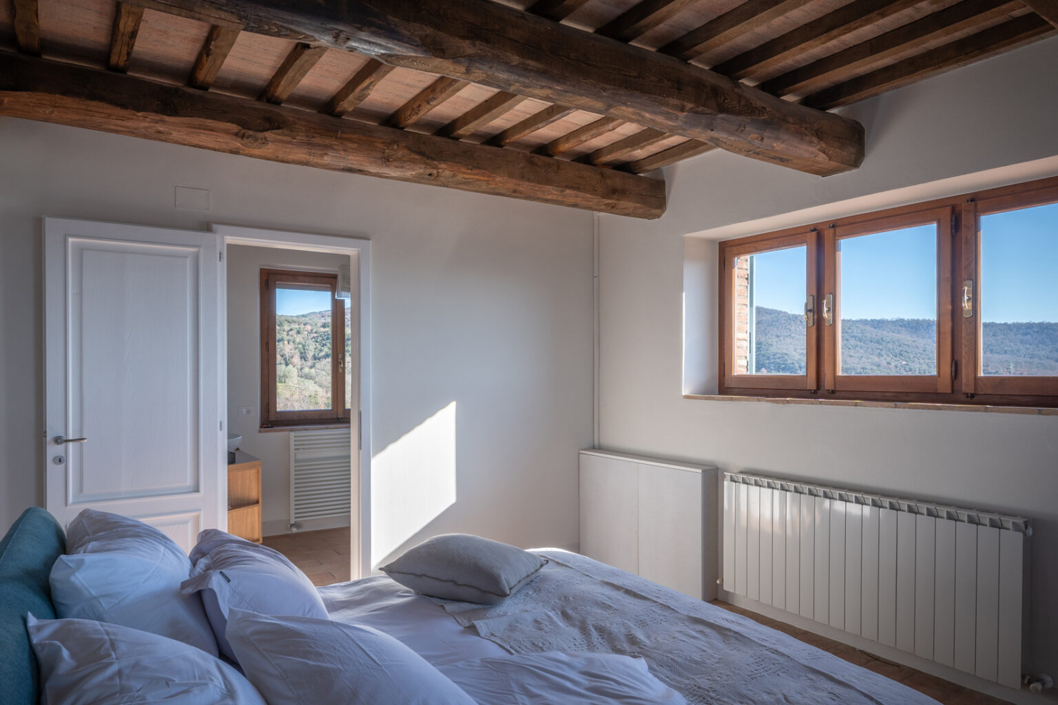 Bright and airy bedroom featuring a cozy bed with white pillows, wooden beams on the ceiling, and large windows offering scenic mountain views. The room includes a radiator and a doorway leading to another space, enhancing the open and inviting atmosphere. | Only on SopranoVillas
