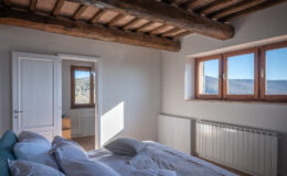 Bright and airy bedroom featuring a cozy bed with white pillows, wooden beams on the ceiling, and large windows offering scenic mountain views. The room includes a radiator and a doorway leading to another space, enhancing the open and inviting atmosphere.