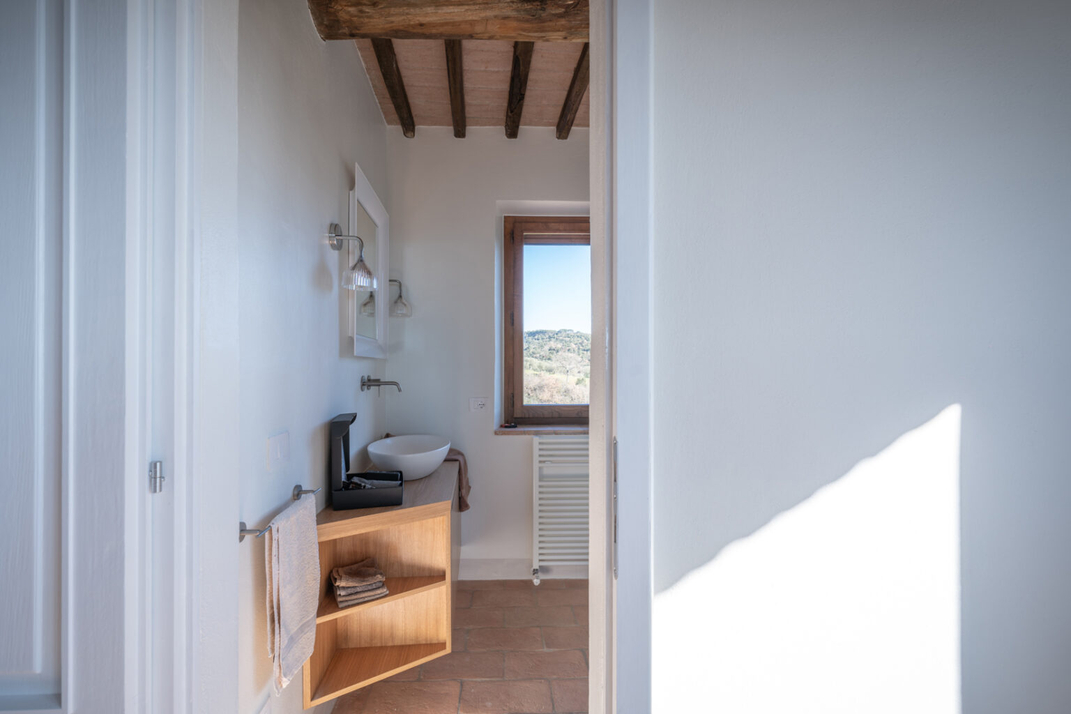 Modern bathroom interior featuring a wooden vanity with a round sink, natural light from a window with a scenic view, and stylish lighting fixtures, showcasing a blend of contemporary and rustic design elements. | Only on SopranoVillas