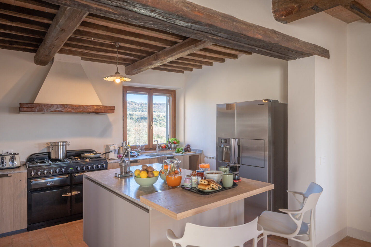 Modern kitchen with rustic wooden beams, featuring a gas stove, stainless steel refrigerator, and a large island with a variety of fresh fruits, baked goods, and beverages, overlooking a scenic view through large windows. | Only on SopranoVillas