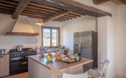 Modern kitchen with rustic wooden beams, featuring a gas stove, stainless steel refrigerator, and a large island with a variety of fresh fruits, baked goods, and beverages, overlooking a scenic view through large windows.