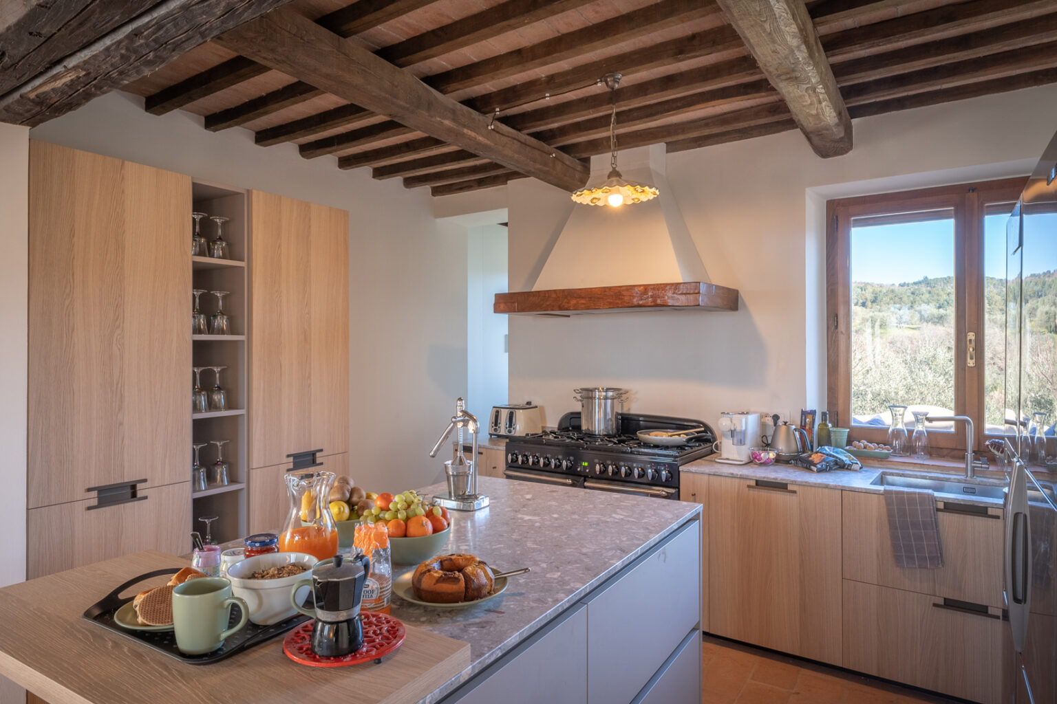 Modern kitchen featuring wooden beams, a spacious countertop with breakfast items, and a scenic view through the window. The kitchen is equipped with stainless steel appliances, a gas stove, and elegant cabinetry, creating a warm and inviting atmosphere. | Only on SopranoVillas