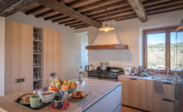 Modern kitchen featuring wooden beams, a spacious countertop with breakfast items, and a scenic view through the window. The kitchen is equipped with stainless steel appliances, a gas stove, and elegant cabinetry, creating a warm and inviting atmosphere.