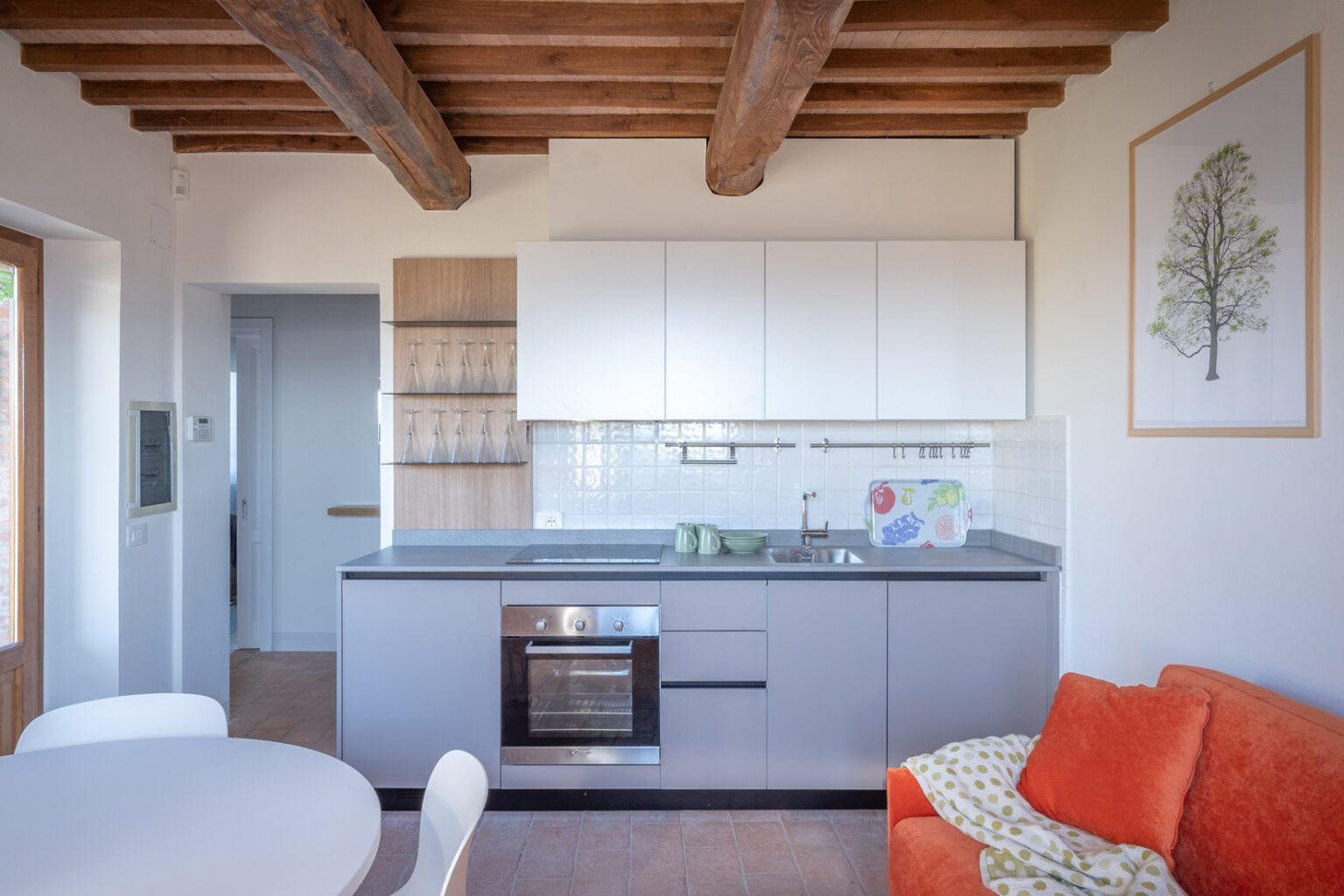 Modern kitchen interior featuring white cabinets, a gray countertop, and wooden beams. The kitchen includes a stainless steel oven, sink, and glassware display. A round dining table and orange sofa add a cozy touch. Natural light enhances the inviting atmosphere. | Only on SopranoVillas