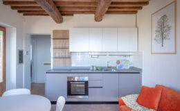 Modern kitchen interior featuring white cabinets, a gray countertop, and wooden beams. The kitchen includes a stainless steel oven, sink, and glassware display. A round dining table and orange sofa add a cozy touch. Natural light enhances the inviting atmosphere.