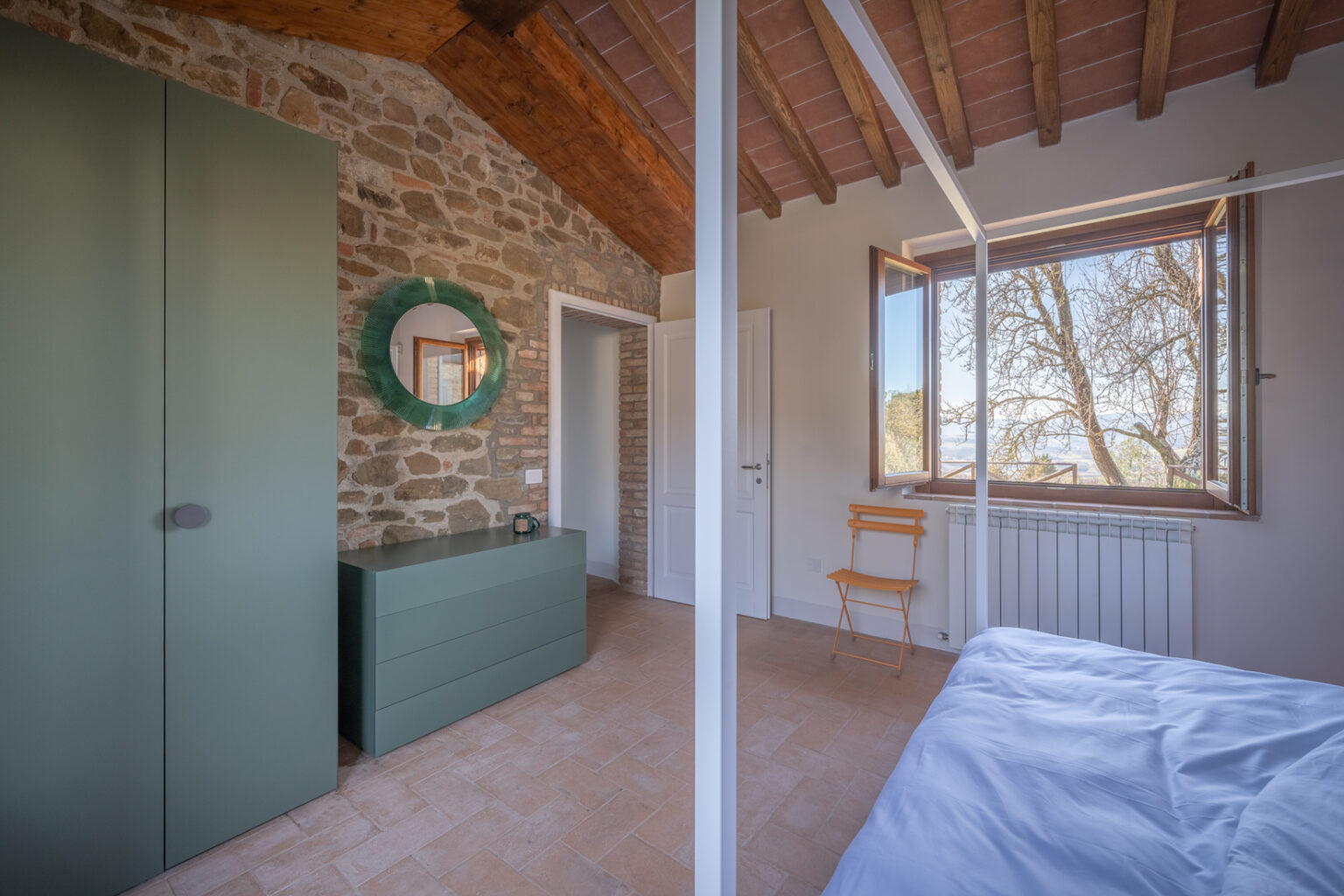 Modern bedroom interior featuring a stone wall, green furniture, a round mirror, and a large window with a scenic view. The room has wooden beams, a cozy atmosphere, and minimalist design elements, including a yellow folding chair and a neatly made bed. | Only on SopranoVillas