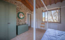 Modern bedroom interior featuring a stone wall, green furniture, a round mirror, and a large window with a scenic view. The room has wooden beams, a cozy atmosphere, and minimalist design elements, including a yellow folding chair and a neatly made bed.