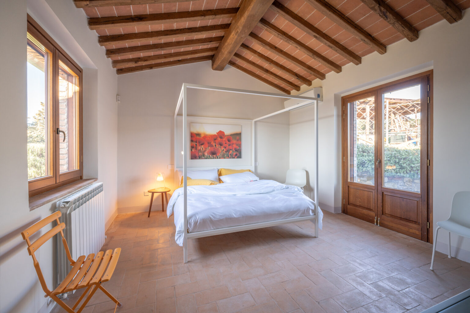 Bright and airy bedroom featuring a four-poster bed with white linens, wooden beams on the ceiling, a cozy bedside lamp, and a large window overlooking a serene outdoor view. The room has a warm, inviting atmosphere with terracotta floors and minimalist decor. | Only on SopranoVillas