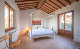 Bright and airy bedroom featuring a four-poster bed with white linens, wooden beams on the ceiling, a cozy bedside lamp, and a large window overlooking a serene outdoor view. The room has a warm, inviting atmosphere with terracotta floors and minimalist decor.