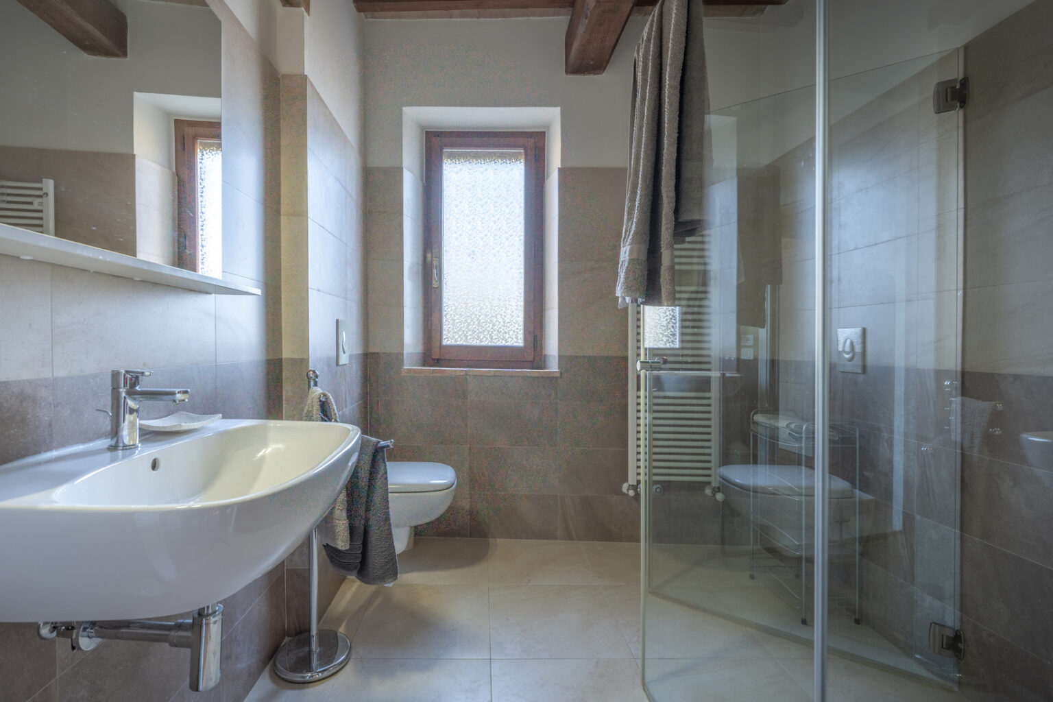 Modern bathroom interior featuring a sleek sink, toilet, and glass shower enclosure. The design includes natural light from a window, warm wood accents, and a towel rack, showcasing a stylish and functional space. | Only on SopranoVillas
