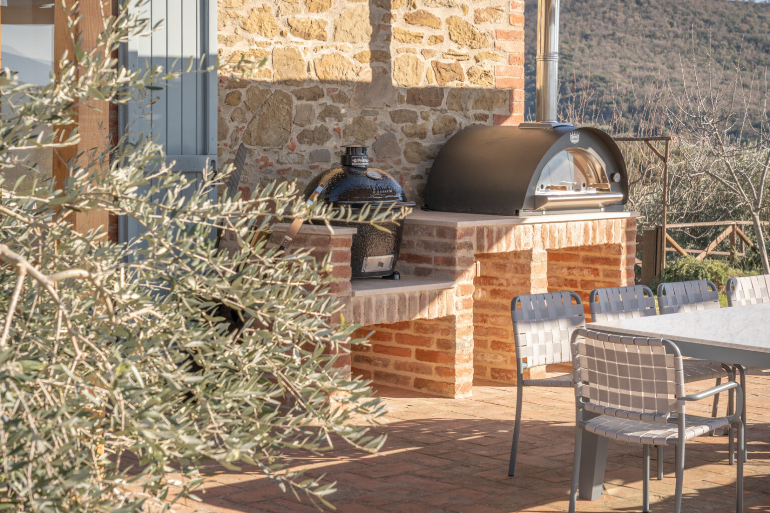 Outdoor kitchen featuring a black ceramic grill and a stainless steel pizza oven on a brick countertop, surrounded by olive branches and a dining table with modern chairs, set against a scenic hillside backdrop. | Only on SopranoVillas