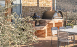 Outdoor kitchen featuring a black ceramic grill and a stainless steel pizza oven on a brick countertop, surrounded by olive branches and a dining table with modern chairs, set against a scenic hillside backdrop.