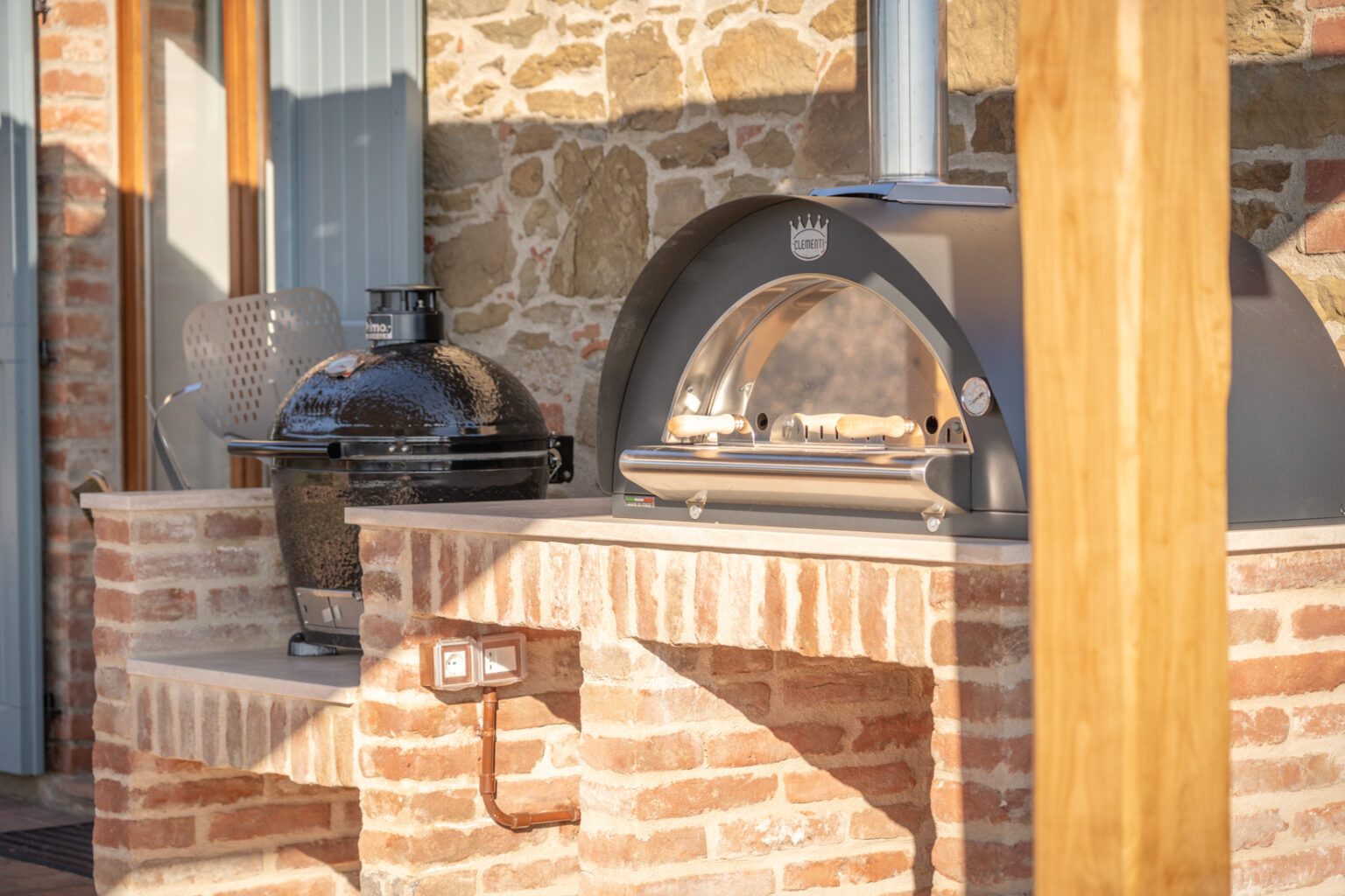 Outdoor kitchen featuring a black ceramic grill and a modern pizza oven, both on a brick countertop, with a rustic stone wall in the background. Ideal for al fresco dining and cooking enthusiasts. | Only on SopranoVillas
