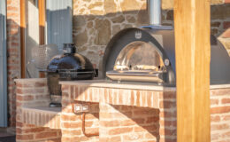 Outdoor kitchen featuring a black ceramic grill and a modern pizza oven, both on a brick countertop, with a rustic stone wall in the background. Ideal for al fresco dining and cooking enthusiasts.