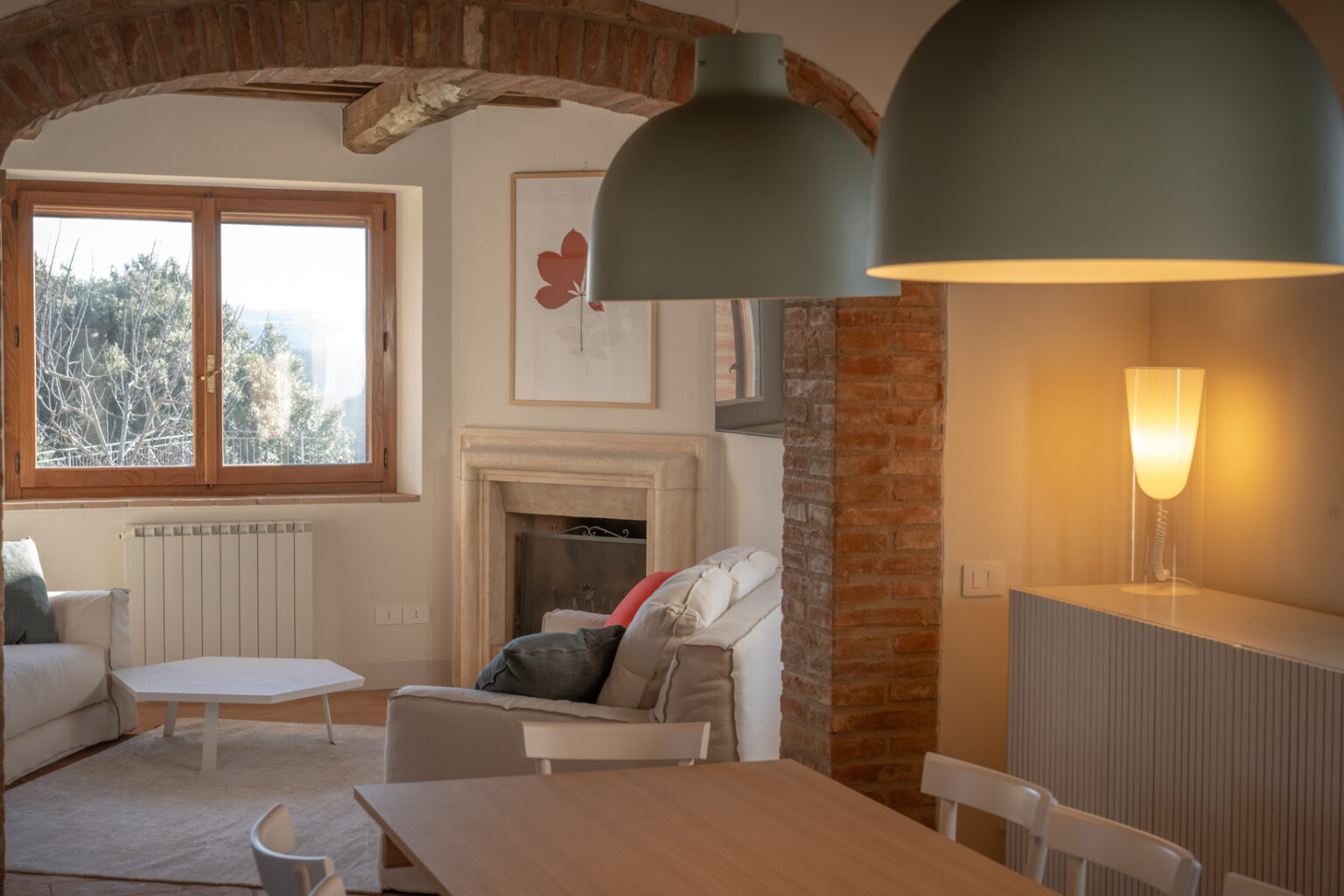 Cozy living room with a modern design featuring a white sofa, a minimalist coffee table, and a warm color palette. Natural light streams through large wooden windows, highlighting a stylish lamp and a classic fireplace. Exposed brick archways add character to the space, creating a welcoming atmosphere. | Only on SopranoVillas