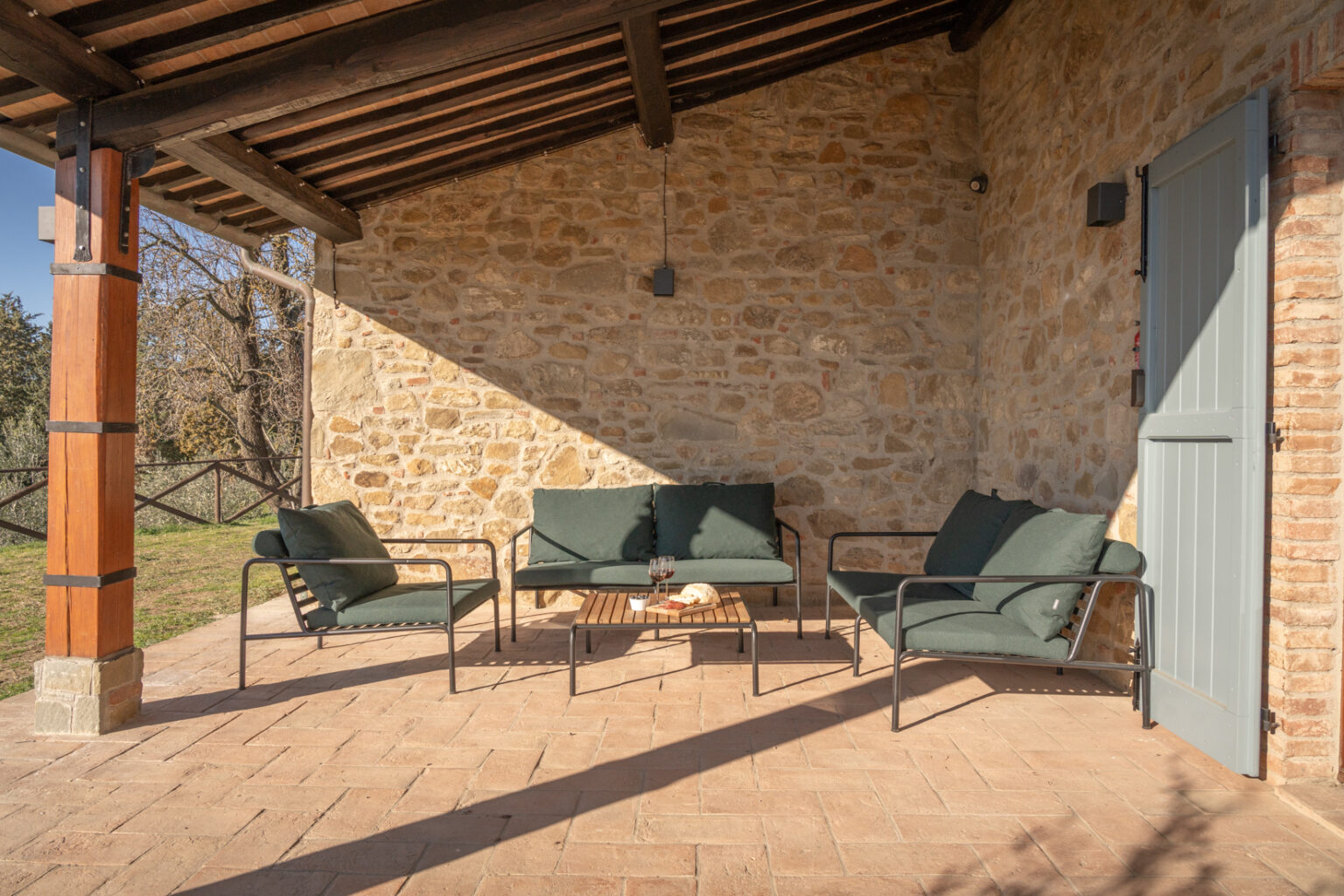 Cozy outdoor seating area featuring green cushions on metal-framed sofas and a wooden coffee table, set against a rustic stone wall and a wooden beam ceiling, perfect for relaxation and enjoying nature. | Only on SopranoVillas