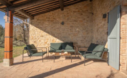 Cozy outdoor seating area featuring green cushions on metal-framed sofas and a wooden coffee table, set against a rustic stone wall and a wooden beam ceiling, perfect for relaxation and enjoying nature.