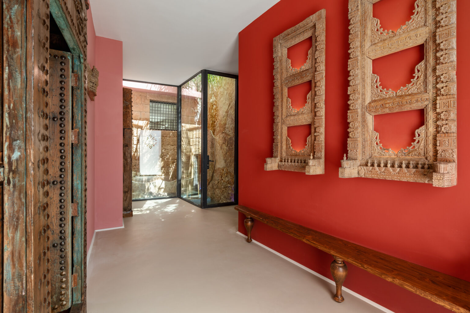 Interior view of a stylish hallway featuring vibrant red walls, intricate wooden wall art, and a sleek glass partition, complemented by a wooden bench. Ideal for showcasing modern design and artistic decor. | Only on SopranoVillas