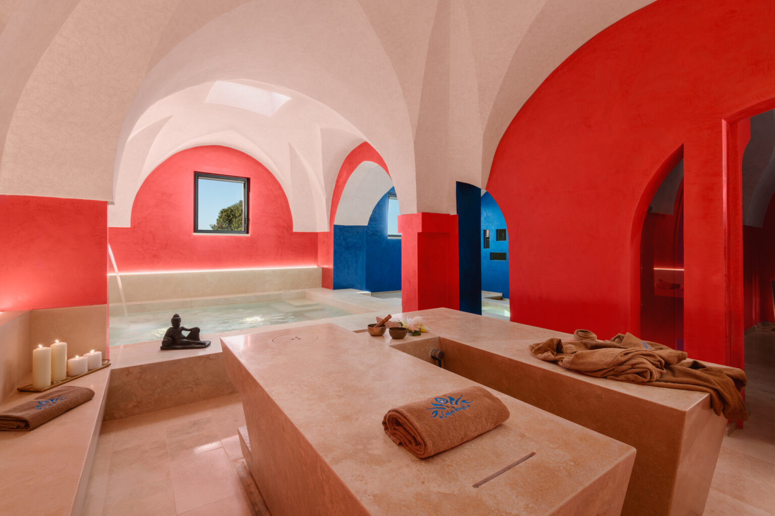 Luxurious spa interior featuring a serene pool, vibrant red and blue walls, and elegant arching ceilings. The space includes candles and soft towels, creating a tranquil atmosphere for relaxation and wellness. | Only on SopranoVillas