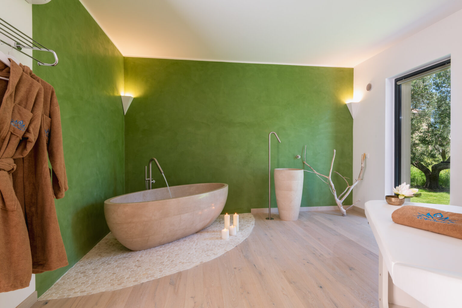 Modern spa bathroom featuring a freestanding wooden bathtub, a minimalist sink, and a vibrant green accent wall. Soft lighting creates a serene atmosphere, complemented by natural elements like candles and a robe hanging on a rack. Large windows offer views of the outdoors. | Only on SopranoVillas
